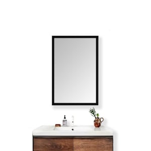 Puede incluir: Un espejo rectangular con marco negro está montado sobre un mueble de baño. El mueble tiene una encimera blanca, un mueble de madera y un lavabo. Una pequeña planta en un jarrón decorativo se encuentra sobre la encimera.