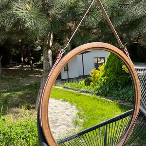 Może przedstawiać: Okrągłe lustro w drewnianej ramie z brązowym sk&oacute;rzanym paskiem do zawieszenia. Lustro odbija zielony trawnik i piaszczystą ścieżkę.