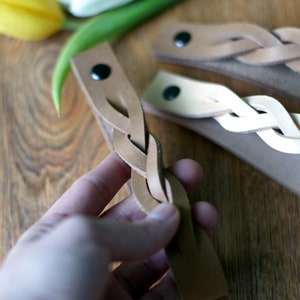 May include: Three braided leather bracelets in different shades of brown. The bracelets are made of thin leather strips that are woven together to create a braided design. The bracelets are shown on a wooden surface.