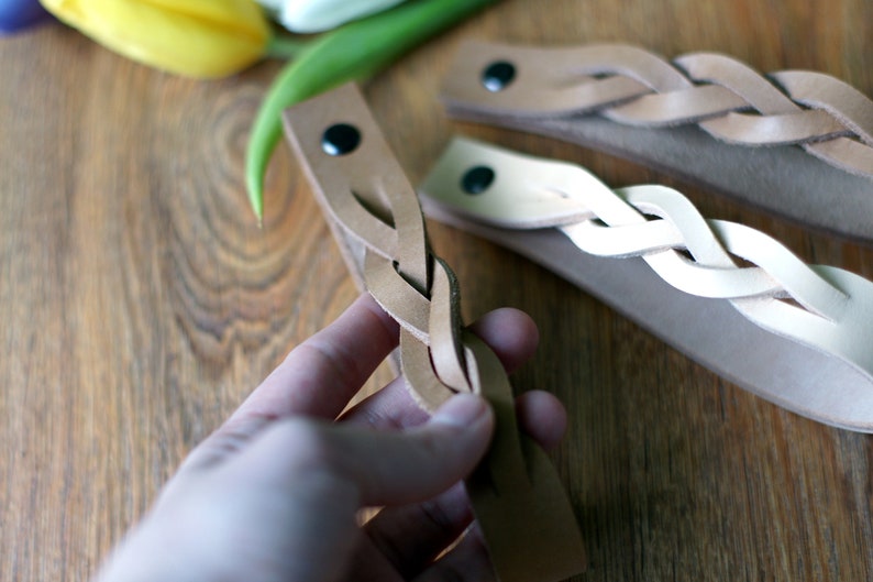 May include: Three braided leather bracelets in different shades of brown. The bracelets are made of thin leather strips that are woven together to create a braided design. The bracelets are shown on a wooden surface.