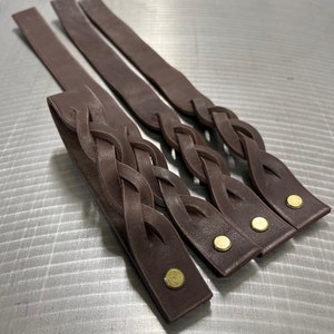 May include: Three brown leather braided straps with brass rivets. The straps are woven together in a complex pattern.