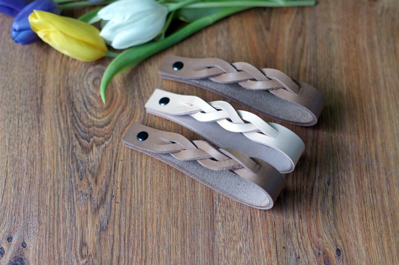 May include: Three braided leather bracelets in brown, beige, and white. The bracelets are on a wooden surface.