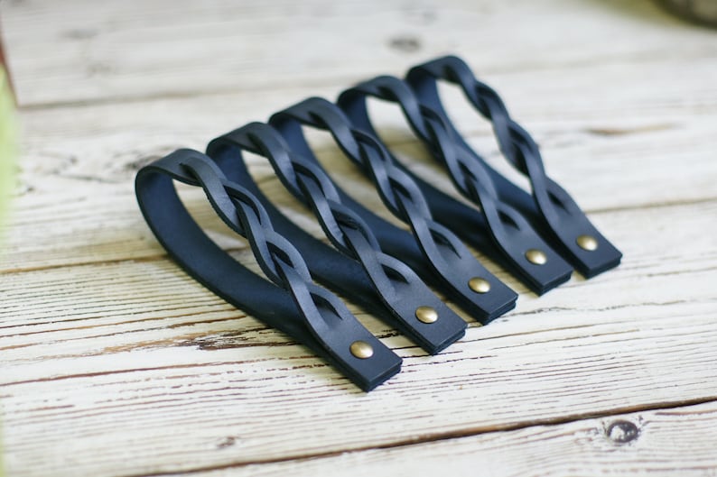 May include: Five dark blue leather braided handles with brass rivets. The handles are arranged in a row on a white wooden surface.