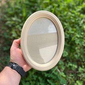 May include: Oval-shaped, cream-colored picture frame. The frame has a smooth, rounded edge and a neutral-toned backing. The frame is held against a blurred green background, suggesting an outdoor setting. The frame is empty, ready for a photo.