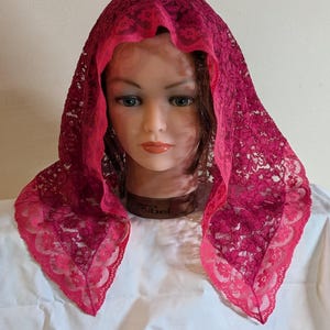 May include: A triangular head covering made of deep magenta lace with a floral pattern and a scalloped edge. The lace features a delicate, open-weave design that drapes over the head, showcasing the intricate floral motifs throughout the fabric.
