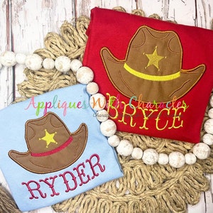 Puede incluir: Dos camisetas infantiles, una roja y otra azul claro, cada una con un diseño de sombrero de vaquero bordado y una estrella dorada. La camiseta roja tiene el nombre "BRYCE" bordado en amarillo, mientras que la azul tiene "RYDER" en rosa.