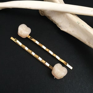 Può includere: Due mollette per capelli dorate con un ornamento in resina bianca a forma di dente su ciascuna estremità.