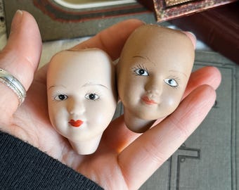 May include: Two antique doll heads with painted features. One head is a pale porcelain color, the other is a tan color. Both have painted blue eyes, thin eyebrows, and red lips. The doll heads are held in a hand.