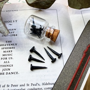May include: A small glass bottle with a cork stopper filled with black tacks. Five black tacks are scattered on a white surface with text. The text reads "The Heavenly Spheres Make Music For Us. All Things Join The Dance."