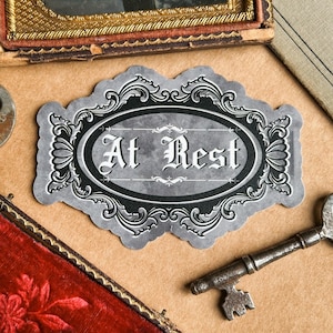 May include: A decorative label with the words "At Rest" in an ornate font. The label is gray with a black oval border and decorative flourishes. A vintage key and other antique items are in the background.