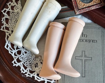 May include: Four porcelain doll legs in two pairs. Two are off-white, and two are a flesh tone. The legs are arranged on a wooden surface with a lace doily and a book with the words "In Memoriam" and a cross.