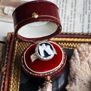 Op de afbeelding: Een zilveren ring met een gehamerde band en een cirkelvormige zwart-witte steen, gepresenteerd in een vintage rode leren ringdoos. De steen van de ring heeft een wit ontwerp op een zwarte achtergrond. De doos is open en toont een rode fluwelen binnenkant.