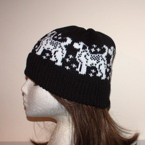 May include: Black knitted beanie with a white pattern of dogs. The beanie is on a white mannequin head with brown hair.