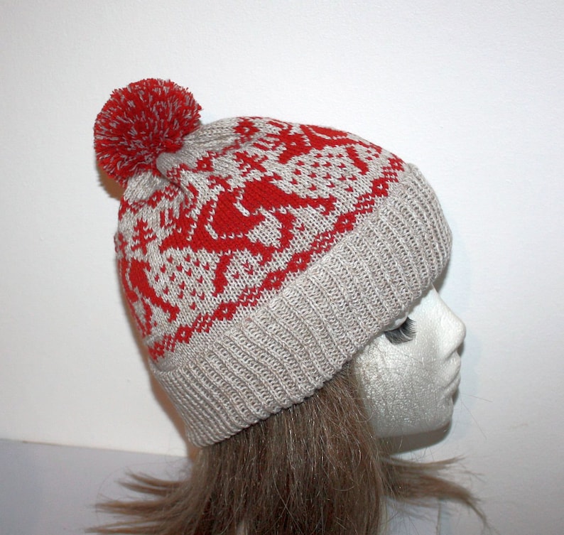 May include: A knitted winter hat with a red and white pattern featuring reindeer and snowflakes. The hat has a ribbed brim and a red pom-pom on top. The hat is displayed on a mannequin head.