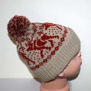 May include: A knitted winter hat with a red and beige pattern featuring reindeer and geometric shapes. The hat has a ribbed brim and a large red pom-pom on top. The hat is designed for cold weather.