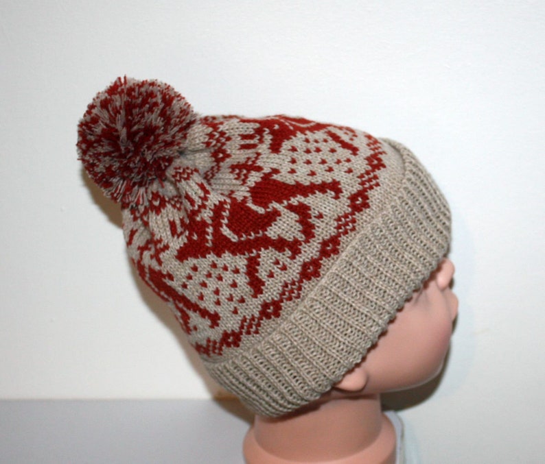 May include: A knitted winter hat with a red and beige pattern featuring reindeer and geometric shapes. The hat has a ribbed brim and a large red pom-pom on top. The hat is designed for cold weather.