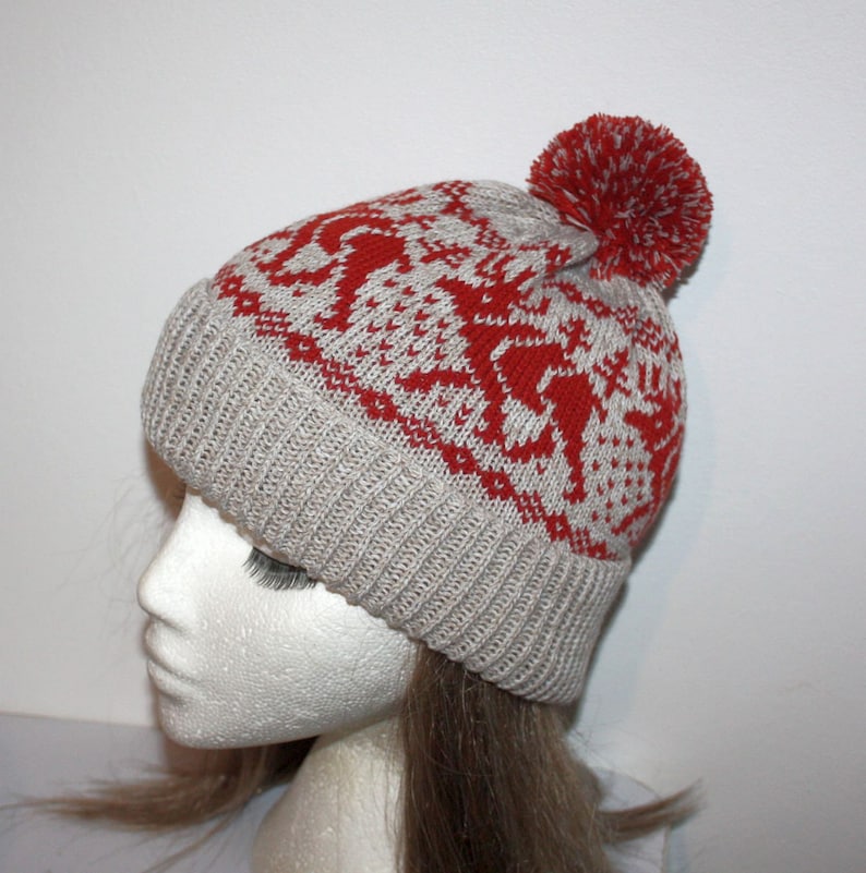 May include: A knitted winter hat with a red and white pattern and a red pom-pom. The hat is light gray with a ribbed brim and a repeating pattern of red shapes. The pom-pom is a solid red color.