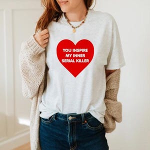 May include: A light gray t-shirt with a large red heart graphic. The heart contains the white text: "YOU INSPIRE MY INNER SERIAL KILLER". The shirt is paired with blue jeans.
