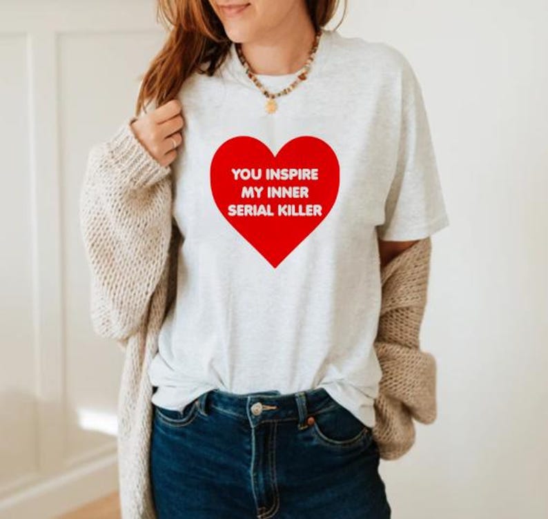 May include: A light gray t-shirt with a large red heart graphic. The heart contains the white text: "YOU INSPIRE MY INNER SERIAL KILLER". The shirt is paired with blue jeans.