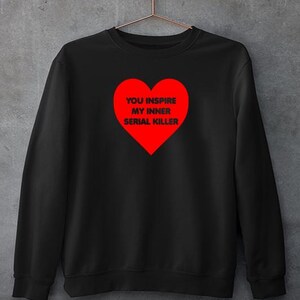 May include: Black sweatshirt with a red heart graphic. The heart contains the text "YOU INSPIRE MY INNER SERIAL KILLER" in white. The sweatshirt is hanging on a metal hanger against a gray background.