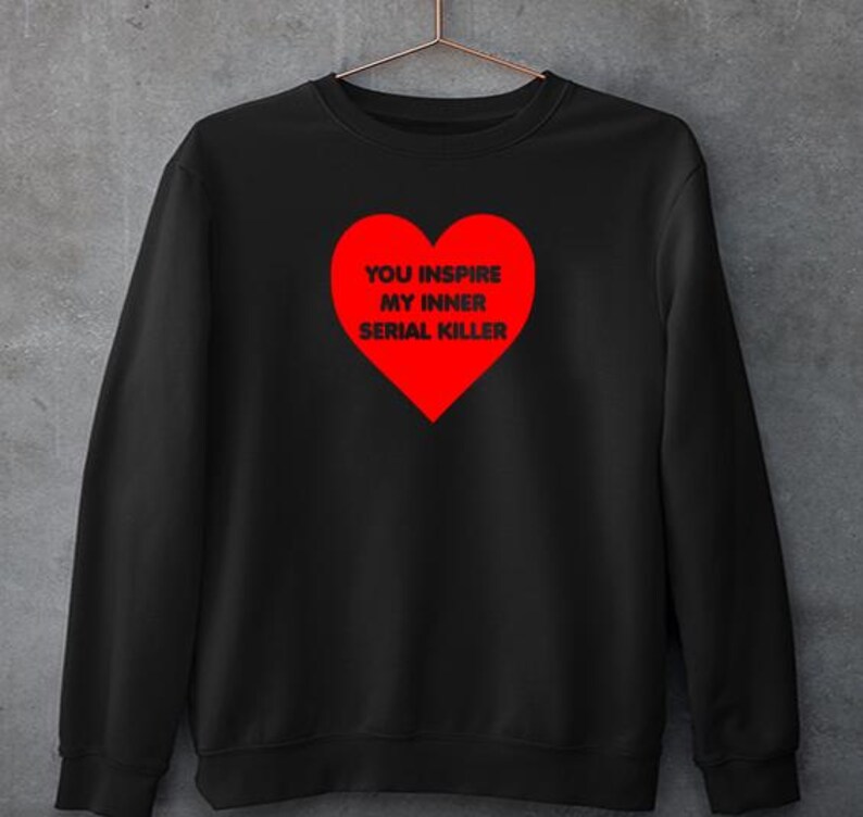 May include: Black sweatshirt with a red heart graphic. The heart contains the text "YOU INSPIRE MY INNER SERIAL KILLER" in white. The sweatshirt is hanging on a metal hanger against a gray background.