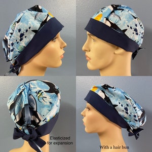 May include: A blue and white patterned surgical scrub hat with a navy blue elastic band and tie. The hat features a design of orcas, fish, and seaweed. The hat is shown on a mannequin head, with the elastic band pulled back to show the expansion of the hat. The text "Elasticized for expansion" is written below the hat. The hat is also shown on a mannequin head with the elastic band tied in a bun. The text "With a hair bun" is written below the hat.