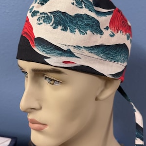 May include: A fabric cap featuring a Japanese wave pattern in black, white, and teal, with red accents. The cap is tied at the back. The design is inspired by traditional Japanese art.