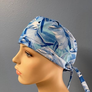 May include: A blue and white patterned scrub cap featuring a repeating dolphin design. The cap is tied at the back and is made of a lightweight fabric. The dolphins are in shades of blue and white.