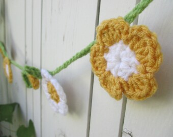 Op de afbeelding: Een gehaakte slinger met gele en witte bloemen en groene bladeren. De slinger hangt aan een wit houten hek.