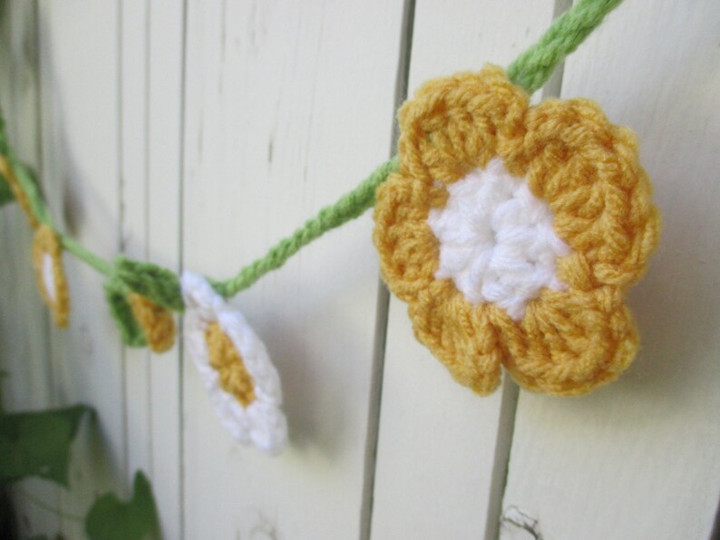 May include: A crocheted garland featuring yellow and white flowers and green leaves. The garland is hanging on a white wooden fence.