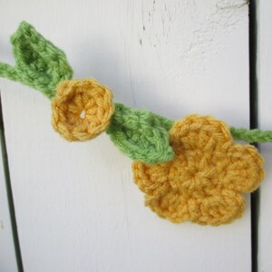 May include: A crocheted garland featuring yellow flowers and green leaves. The garland is hanging on a white wooden surface.