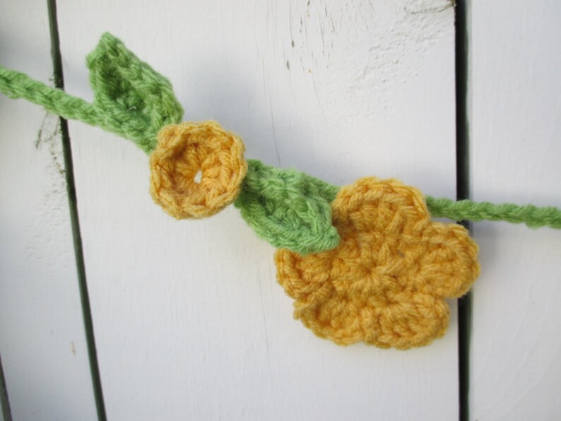 May include: A crocheted garland featuring yellow flowers and green leaves. The garland is hanging on a white wooden surface.