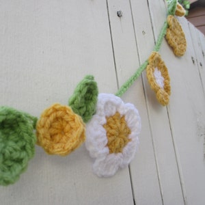 May include: A crocheted garland featuring white, yellow and green flowers. The garland is hanging on a white wooden surface.