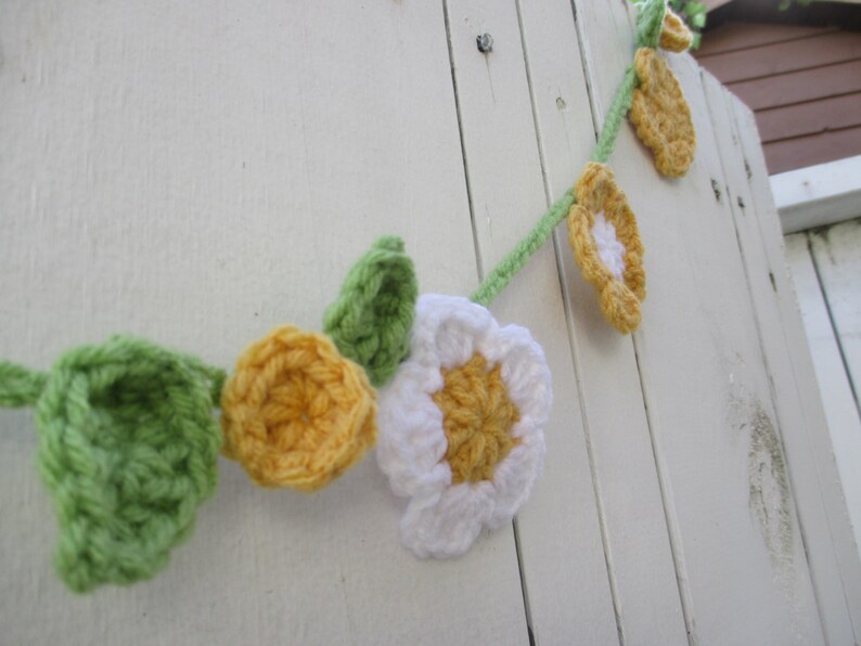 May include: A crocheted garland featuring white, yellow and green flowers. The garland is hanging on a white wooden surface.
