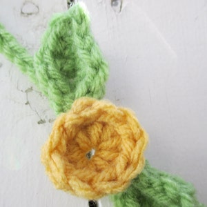 May include: A crocheted garland featuring yellow flowers and green leaves. The garland is made from yarn and is ideal for decorating a home or party.
