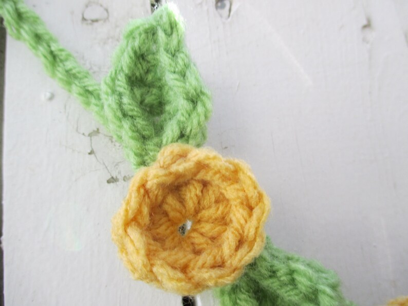 May include: A crocheted garland featuring yellow flowers and green leaves. The garland is made from yarn and is ideal for decorating a home or party.