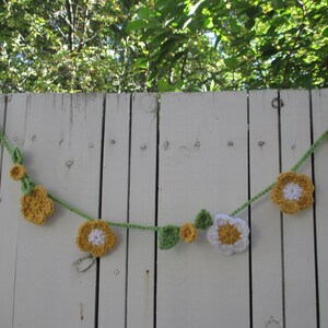 May include: A crocheted garland with white and yellow flowers and green leaves hanging on a white wooden fence.