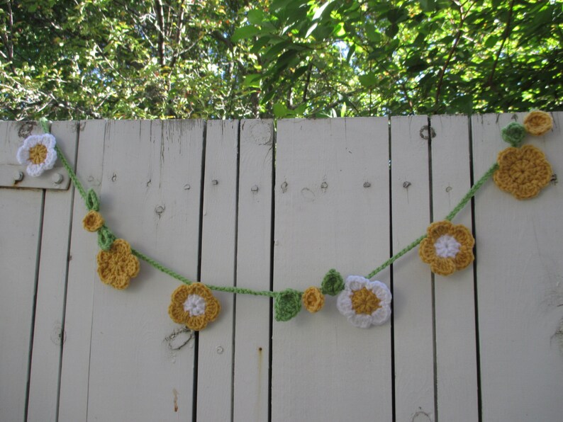 May include: A crocheted garland with white and yellow flowers and green leaves hanging on a white wooden fence.