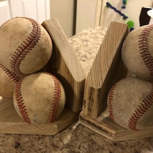 May include: Baseball-themed bookends. Each bookend is made of light-colored wood and features a baseball. The baseballs are tan with red stitching. The bookends are designed to hold books upright.
