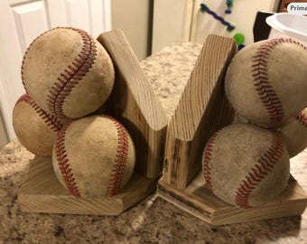 May include: Baseball-themed bookends. Each bookend is made of light-colored wood and features a baseball. The baseballs are tan with red stitching. The bookends are designed to hold books upright.