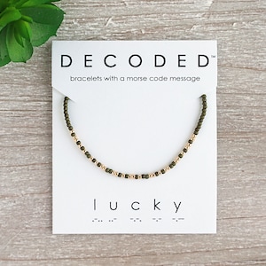May include: A green and gold beaded bracelet with the word "lucky" and its Morse code representation on a white card. The card also has the brand name "DECODED" and the tagline "bracelets with a morse code message".