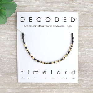 May include: Black and gold beaded bracelet with the word "timelord" and its morse code translation on a white card. DECODED™ bracelets with a morse code message.