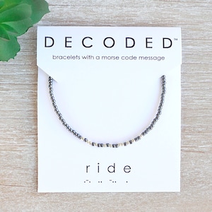 May include: A beaded bracelet with a Morse code message on a white card. The card reads "DECODED" and "ride" in a minimalist font. The bracelet is dark gray with gold accents. The background is a light wood grain.