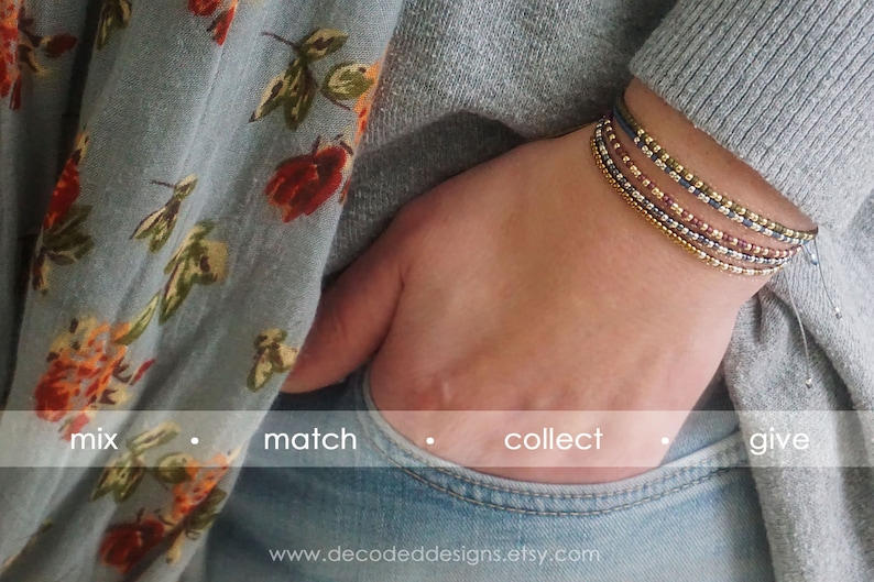 May include: A close-up of a person's wrist wearing a stack of colorful beaded bracelets. The bracelets are made of small, round beads in shades of gold, silver, red, and blue. The bracelets are layered on top of each other, creating a unique and stylish look. The text "mix match collect give" is visible at the bottom of the image.