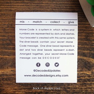 May include: White card with black text explaining how to decode a Morse code bracelet. The text reads "mix match collect give Morse Code is a system in which letters and numbers are represented by dots and dashes. Your bracelet is created with this same system. The silver beads contain your secret Morse Code message. One silver bead represents a dot and two silver beads represent a dash. Arranged together, your secret Morse Code message can be DECODED." The card also includes social media icons and the text "@DecodedUpdates www.decodeddesigns.etsy.com Back of display card."
