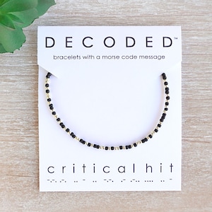May include: Black and gold beaded bracelet with the word "critical hit" and its Morse code translation on a white card. The card has the brand name "DECODED" and the text "bracelets with a morse code message".