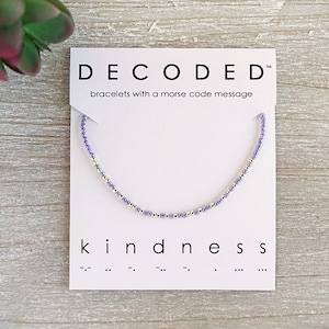 May include: A purple and silver beaded bracelet with the word "kindness" and its Morse code translation on a white card. The card also says "DECODED" and "bracelets with a morse code message".