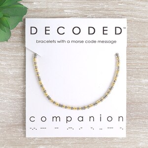 May include: A silver and gold beaded bracelet with the word "companion" and its Morse code translation on a white card. The card also has the text "DECODED" and "bracelets with a morse code message".
