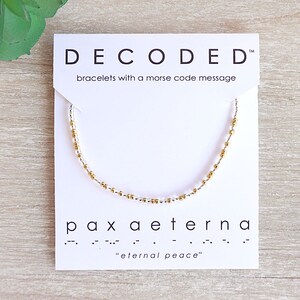 May include: A white card with the text "DECODED" and "bracelets with a morse code message". A gold and white beaded bracelet is displayed on the card. The text "pax aeterna" and "eternal peace" is also on the card.