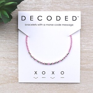 May include: A pink beaded bracelet with a Morse code message, presented on a white card. The card reads "DECODED" and "bracelets with a morse code message". Below the bracelet are the letters "XOXO" in Morse code.
